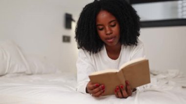 Siyah bir kadın rahat bir şekilde yatağında uzanıyor, kitap okumaya dalmış bir halde. Oda huzurlu ve samimi bir atmosfer yayıyor, rahatlamak ve gevşemek için mükemmel.