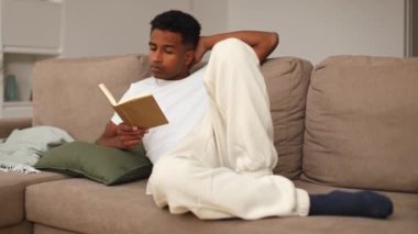 Genç siyahi bir adam bej bir kanepede rahat bir şekilde uzanıyor, bir kitaba dalmış rahat oturma odasında bitkiler ve yumuşak aydınlatma var, rahatlatıcı ve davetkar bir atmosfer yaratıyor.
