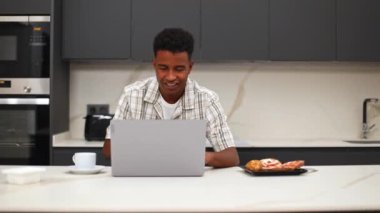 Modern bir mutfakta oturup kahve içerken bilgisayarındaki video görüşmesine konsantre olan siyahi bir adam bir tabak hamur işi yakınlarda, rahat ve üretken bir atmosfer yaratıyor.