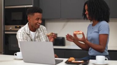 Siyah bir çift, modern mutfaklarında dizüstü bilgisayarlarında bir şeyler izlerken kahvaltıdan zevk alır. Sıcak atmosfer ve rahat hava, birlikte rahatlamış bir sabah rutinini ön plana çıkarır.