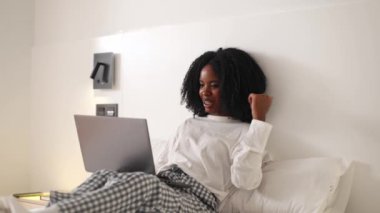 Yatakta siyah bir kadın, dizüstü bilgisayarına gülümseyen bir videoda rahat bir şekilde beyaz bir takım elbise ve ekose pantolon giyiyor, rahat ve üretken bir ortam yaratıyor.