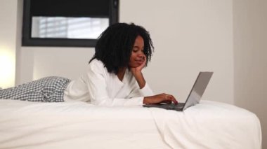 Gündelik giyinmiş siyah bir kadın laptopuyla uğraşırken rahat bir şekilde yatağında yatıyor. Yatak odası konforu, iş ve eğlence için huzurlu bir ortam yaratıyor.