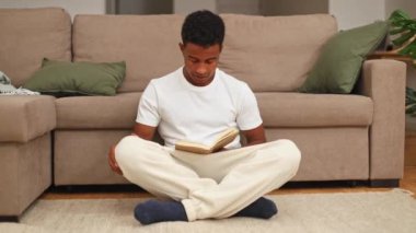 Genç siyah bir adam yerde bağdaş kurup kitap okumaya dalmış, rahat bir bej kanepe ve dekoratif yastıkla rahat bir oturma odasının zemininde oturuyor.