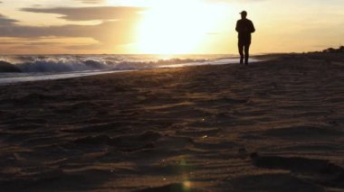 Canlı bir gün batımında kumlu sahilde koşan bir adamın silueti dalgaların ve batan güneşin dinamik hareketlerini yakalıyor..