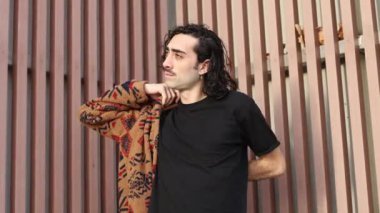 Uzun kıvırcık saçlı ve bıyıklı bir adam güneşli bir günde metal bir çitin önünde dikilirken bronz tenini, desenli ceketini siyah bir gömleğin üzerine yerleştirir.