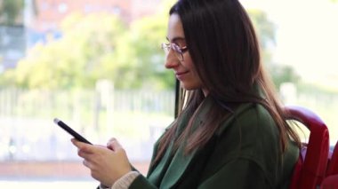 Yeşil ceketli bir kadın otobüste akıllı telefonunu kullanırken gülümsüyor. Güneş ışığı pencereden süzülüyor, içerik ifadesini yazarken aydınlatıyor.