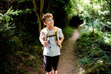 Gözlüklü ve sırt çantalı neşeli genç bir çocuk Canfranc, Huesca, İspanya 'da yemyeşil bir orman yolunda yürürken gülümsüyor.