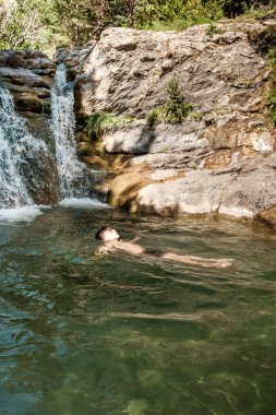 Canfranc, Huesca, İspanya 'da engebeli kayalar ve yemyeşil yeşilliklerle çevrili mütevazı bir şelalenin altında berrak, doğal bir havuzda yüzen bir adamı gösteren sakin bir görüntü.