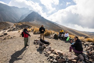 Bir grup yürüyüşçü, arka planda dramatik dağ zirveleri ve parçalı bulutlu gökyüzü olan altın otlarla çevrili kayalık bir volkanik yaylada mola verir..