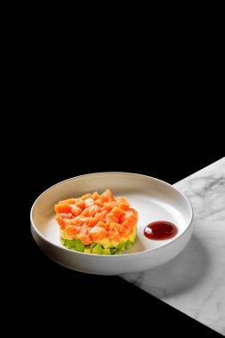 Sos eşliğinde beyaz bir tabakta servis edilen doğranmış avokado ile sunulmuş güzel bir somon tartare, siyah ve mermer bir zemin üzerine kurulmuş, modern bir estetik yaratıyor.