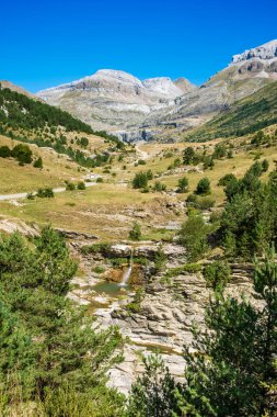 A captivating landscape showcasing the rugged beauty of Canfranc in Huesca, Spain, featuring majestic mountains and a serene cascading waterfall amid lush greenery clipart