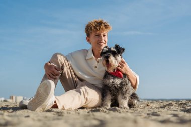 Genç bir insan kumsalda güneşli bir günün tadını çıkarır tüylü yoldaşlarıyla kumda otururken, yukarıdaki açık mavi gökyüzü ile neşeli bir anı paylaşırlar.