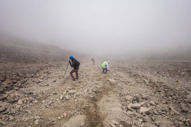 Bir grup yürüyüşçü yoğun siste dik, kayalık bir yamaçtan inerek, zorlu koşullarda dayanıklılık ve kararlılık gösteren, yürüyüş direkleriyle engebeli volkanik arazide yol alırlar..
