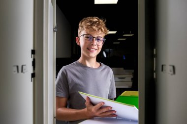 Gözlüklü neşeli bir genç çocuk açık bir okul dolabında duruyor, renkli ders kitaplarını tutarken gülümsüyor, pozitif ve çalışkan bir atmosfer sunuyor..