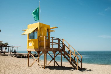 Canlı sarı bir cankurtaran kulesi açık mavi gökyüzünün altında kumlu bir kumsalda duruyor, yeşil bir bayrak dalgalanırken sakin deniz kenarı yaz tatili atmosferini anımsatıyor, kıyı temaları için mükemmel.
