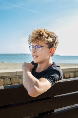 Gözlüklü bir genç, deniz kenarında güneşli bir günün keyfini çıkarır, bir tebessümle bankta otururken okyanus ve mavi gökyüzü huzurlu bir arkaplan sağlar, rahatlatıcı bir anı yakalar.