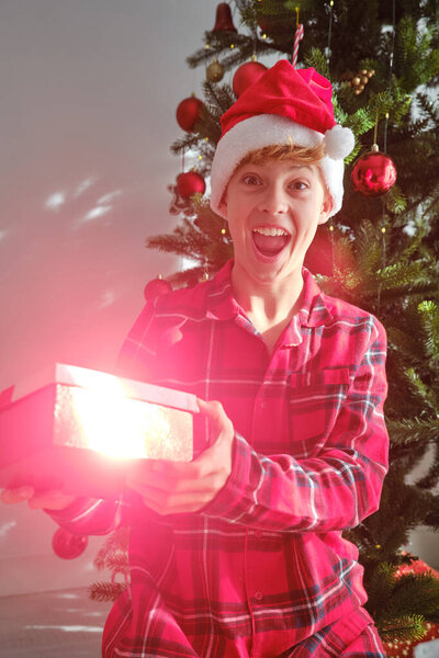 Радостный подросток в шляпе Санты открывает ярко светящийся рождественский подарок возле украшенной елки, выражая удивление и счастье.
