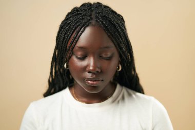 Portrait of young African American female with Afro braids in white pullover standing on beige background in studio clipart