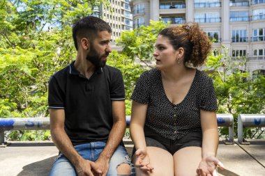Genç bir erkek ve kadın, güneşli bir günde, arka planda yemyeşil ve modern mimariyle birlikte, ciddi bir sohbete dalarlar.