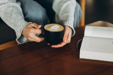 Huzurlu bir an, siyah bir fincanda latte tutarken, ahşap bir masada açık bir kitap, rahatlama ve rahatlığı göstermek için mükemmel.