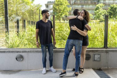 Dışarıda üç arkadaşla neşeli bir buluşma sahnesi. Bir adam, bir çiftin, yemyeşil bir çevre tarafından kuşatılmış güneşli, modern bir kaldırımda sarılışını izlerken gülümsüyor.