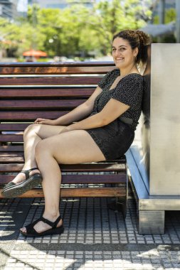 Neşeli genç bir kadın parktaki ahşap bir bankta oturur, polka noktalı bluz ve kot şortla gündelik bir kıyafetle güneşli arka plan ve canlı yeşillik onun neşeli ifadesini güçlendirir.