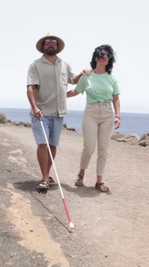 Beyaz bastonlu görme engelli bir adam, güzel bir sahil yolu boyunca bir kadın tarafından yönlendirilir. Her ikisi de sıradan giyinmiş, destekleyici ve huzurlu bir anı tasvir ediyorlar..