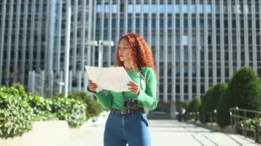 Yeşil kazaklı Latin bir kadın elinde harita tutarken şehir manzarasını keşfediyor. Güneşli bina ve boynundaki kamera seyahat temasını gösteriyor..