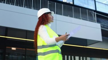 Sert şapka ve güvenlik yeleği giymiş melez bir ırk kadını, şehir ortamında mimari planları inceleyerek, yapı ve şehir gelişiminde çeşitliliği ve ilerlemeyi simgeliyor..