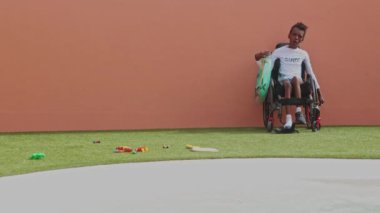 Serebral felçli genç bir zenci çocuk, bir sörf kampında havuz kenarında tekerlekli sandalyede oturuyor, elinde bir şamandıra, etrafı dağınık oyuncaklarla çevrili, eğlenceli bir doğa deneyimi için hazır..