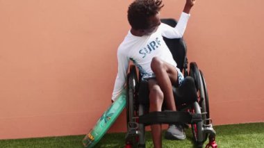 Serebral felçli genç bir zenci çocuk, bir sörf kampında havuz kenarında tekerlekli sandalyede oturuyor, elinde bir şamandıra, etrafı dağınık oyuncaklarla çevrili, eğlenceli bir doğa deneyimi için hazır..