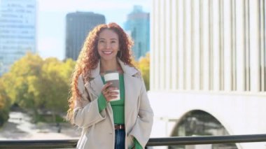 Kıvırcık saçlı melez bir kadın dışarıda kahve fincanı tutuyor. Çağdaş şehir binalarıyla çevrelenmiş bir şehir ortamında neşe ve güven saçıyor..
