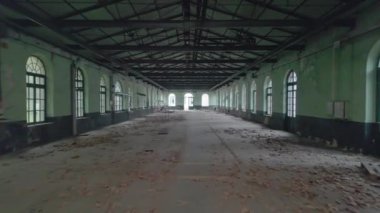 Uzun kemerli pencereler, metal kirişler ve yere saçılmış döküntülerle dolu terk edilmiş bir fabrikanın içi. Çürümüş ayar endüstriyel terk edilmeyi vurguluyor.