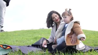 Genç bir aile dışarıda eğleniyor. Anne küçük kızının saçını nazikçe yapıyor, doğal ortamında hassas bir aile etkileşimi sergiliyor..