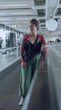 Sırt çantalı gülümseyen bir kadın havaalanı terminalinden geçiyor, yan tarafa bakıyor, muhtemelen uçağa yetişmeye çalışıyor..