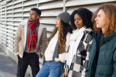 Kış kıyafetleri giyen dört farklı üniversite öğrencisi bir kampüs binasının dış duvarına yaslanıyor.