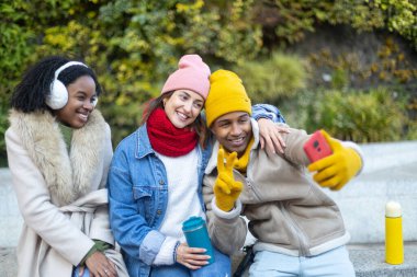 Üç neşeli genç arkadaş, soğuk bir kış gününde, sıcak şapkalar, eşarplar ve eldivenlerle dışarıda selfie çekiyorlar.