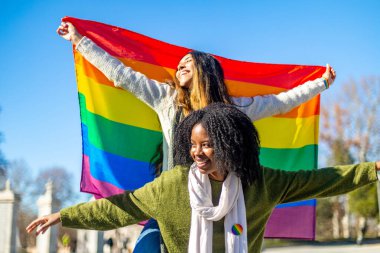 Gökkuşağı bayrağı taşıyan iki neşeli çoklu etnik kadın, güneşli bir günde çeşitliliği ve lgbtq artı hakları kutluyorlar.