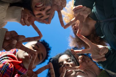 Mavi gökyüzüne karşı parmaklarıyla yıldız şekli oluşturan mutlu çoklu etnik arkadaşlar birliği, takım çalışmasını ve arkadaşlığı simgeliyor.
