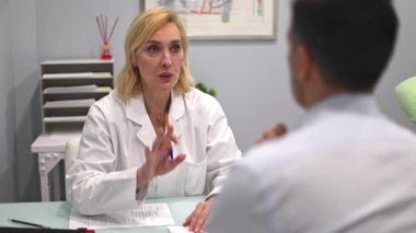 Beyaz önlüklü bir kadın doktor modern bir güzellik kliniğinde erkek bir hastaya danışarak masadaki belgeler üzerinde tedavi seçeneklerini tartışıyor.