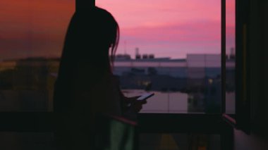Karanlık odada pencerenin yanında duran ve gün batımında pembe bulutlu gökyüzüne karşı tuş takımı ile mesaj gönderen uzun saçlı kadının yavaş görüntüsü.