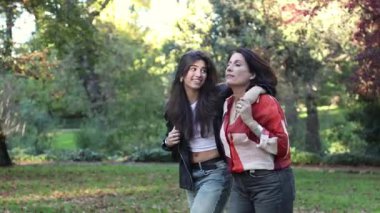 Bir anne ve kızı bir parkta kol kola yürür, neşeli bir sohbete dalarlar. Kızı dinlerken içten bir gülümseme sergiliyor, güçlü bağlarını ve doğadaki bağlantılarını gösteriyor..