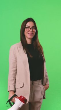 Gözlüklü mutlu bir genç kadın, yeşil arka planda el kamerasıyla konuşurken kameraya bakıyor.