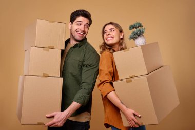 Neşeli genç bir çift, her biri heyecan verici bir hamleye hazırlanırken ellerinde karton kutular olan sırt üstü ayakta tasvir ediliyor. Sıcak ve enerjik arka plan neşe dolu atmosfere ekleniyor.