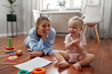 Neşeli bir anne, meraklı bebeğinin hayallerini canlandıran renkli oyuncaklarla çevrili sıcak ahşap bir zeminde canlı çizimler ve eğlenceli aktivitelerle yaratıcılığı keşfetmesini izler.