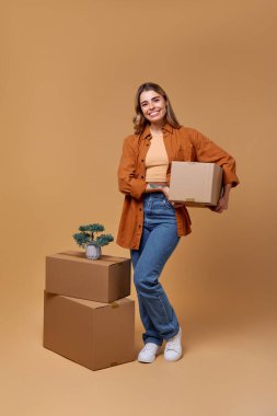 Günlük giysiler içinde neşeli bir kadın elinde çeşitli eşyalarla dolu bir kutu tutarken gülümsüyor. Taşıma malzemeleriyle çevrili. Heyecan verici geçişini ve hayat yolculuğunda yeni başlangıçlarını simgeliyor.