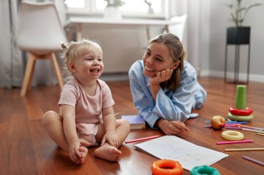 Neşeli bir anne, meraklı bebeğinin hayallerini canlandıran renkli oyuncaklarla çevrili sıcak ahşap bir zeminde canlı çizimler ve eğlenceli aktivitelerle yaratıcılığı keşfetmesini izler.