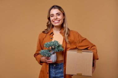 Neşeli genç bir kadın bir elinde canlı bir saksı bitkisi, diğer elinde sağlam bir karton kutu tutuyor. Bu da onun heyecan verici hareketini ve sonsuz olanaklarla dolu yeni bir başlangıcı simgeliyor.