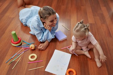 Neşeli bir anne, meraklı bebeğinin hayallerini canlandıran renkli oyuncaklarla çevrili sıcak ahşap bir zeminde canlı çizimler ve eğlenceli aktivitelerle yaratıcılığı keşfetmesini izler.