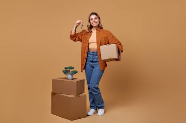 Kutu ve anahtarla neşeli genç bir kadın taşınan kutular ve canlı bir bitki ile çevrili, sıcak bir bej arka plan, somutlaştırıcı yaşam tarzı, yer değiştirme ve yeni başlangıçlar temaları karşı